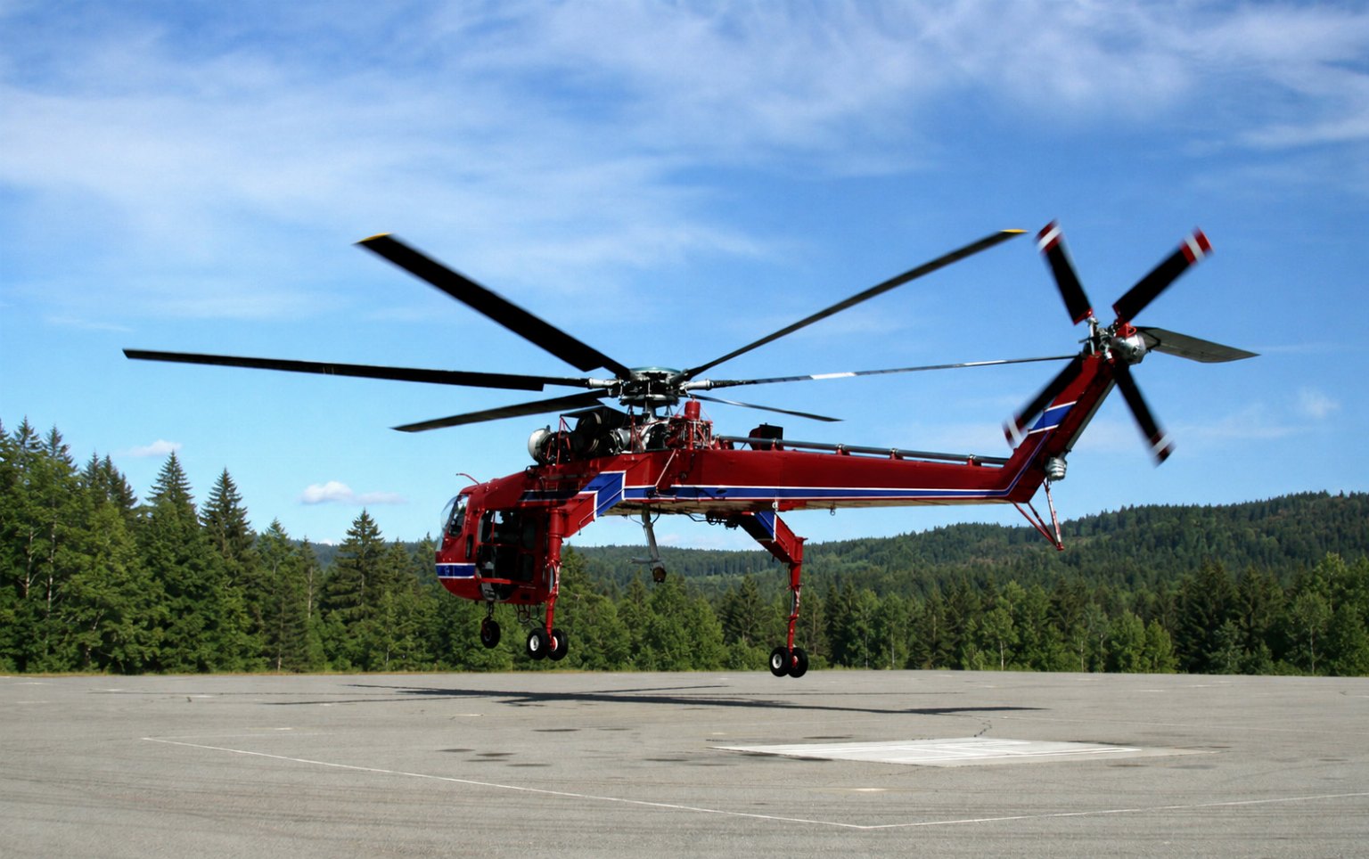 A red Sikorsky S-64 Skycrane just above a paved helipad with landing gear light on the wheels, forest treeline behind