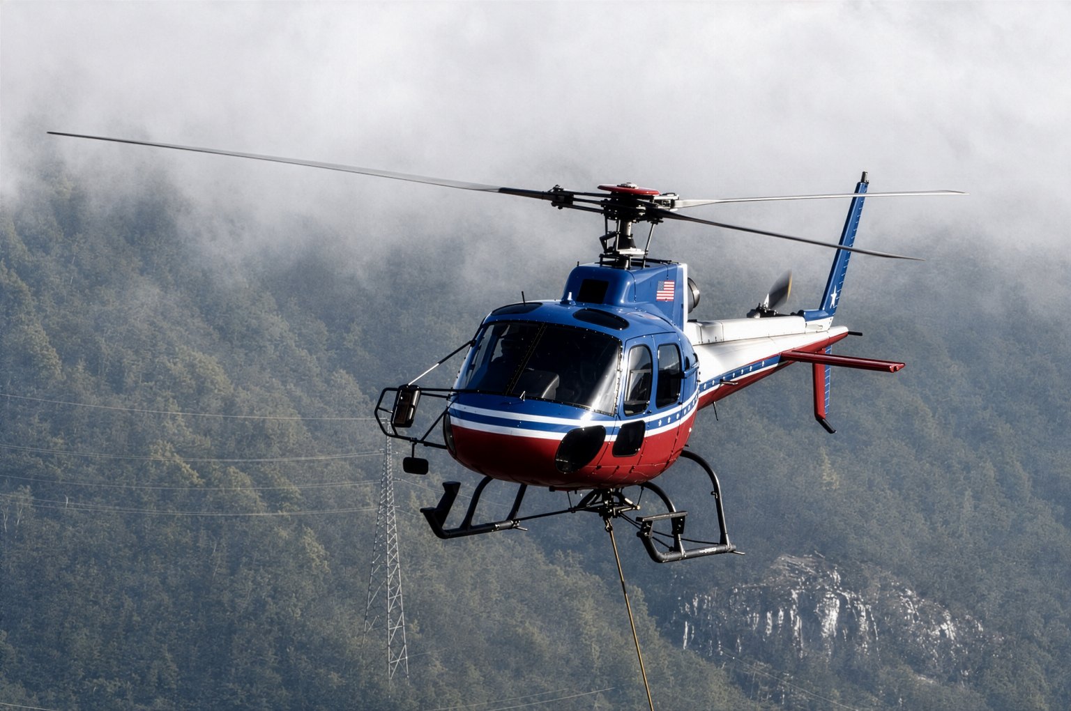 A light single-engine helicopter in a red, white, and blue livery holding a hover with a longline near a transmission tower on a fog-covered mountainside