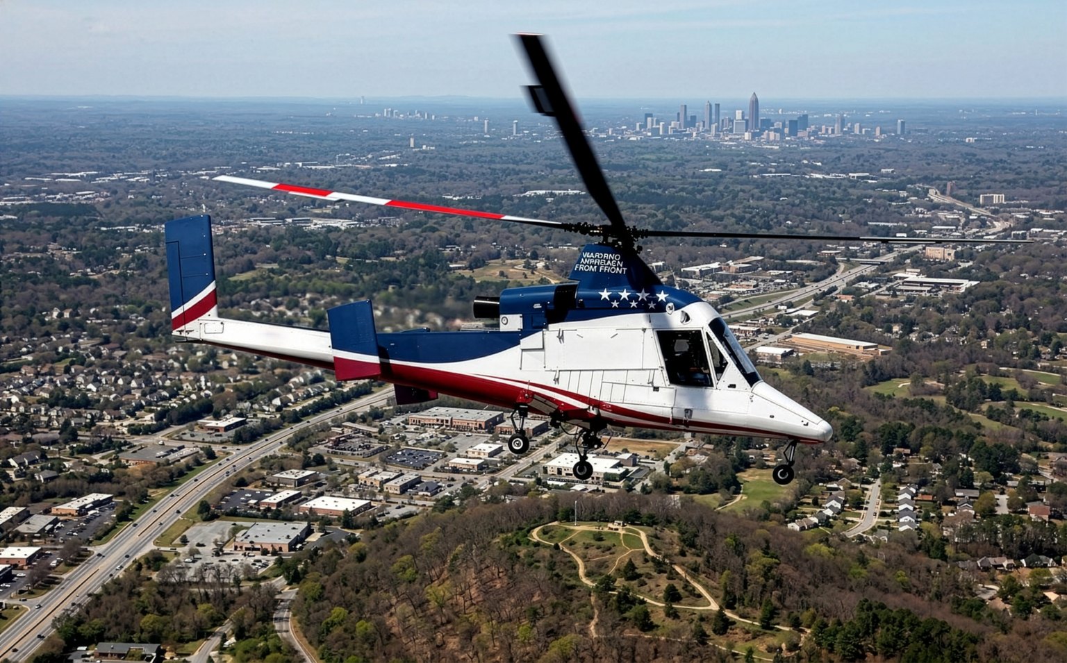 A Kaman K-MAX helicopter in cruising flight above a developed area, with a distant city skyline on the horizon