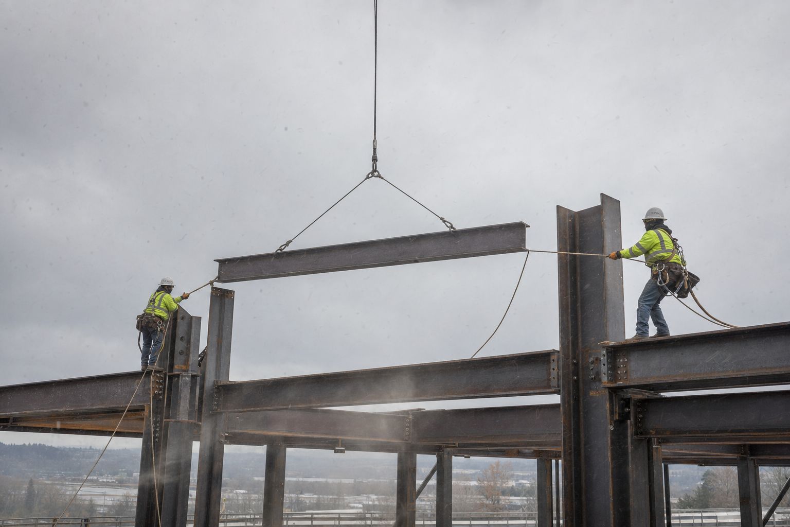 Two ironworkers on open structural steel framing guiding a suspended steel beam into a column connection with tag lines