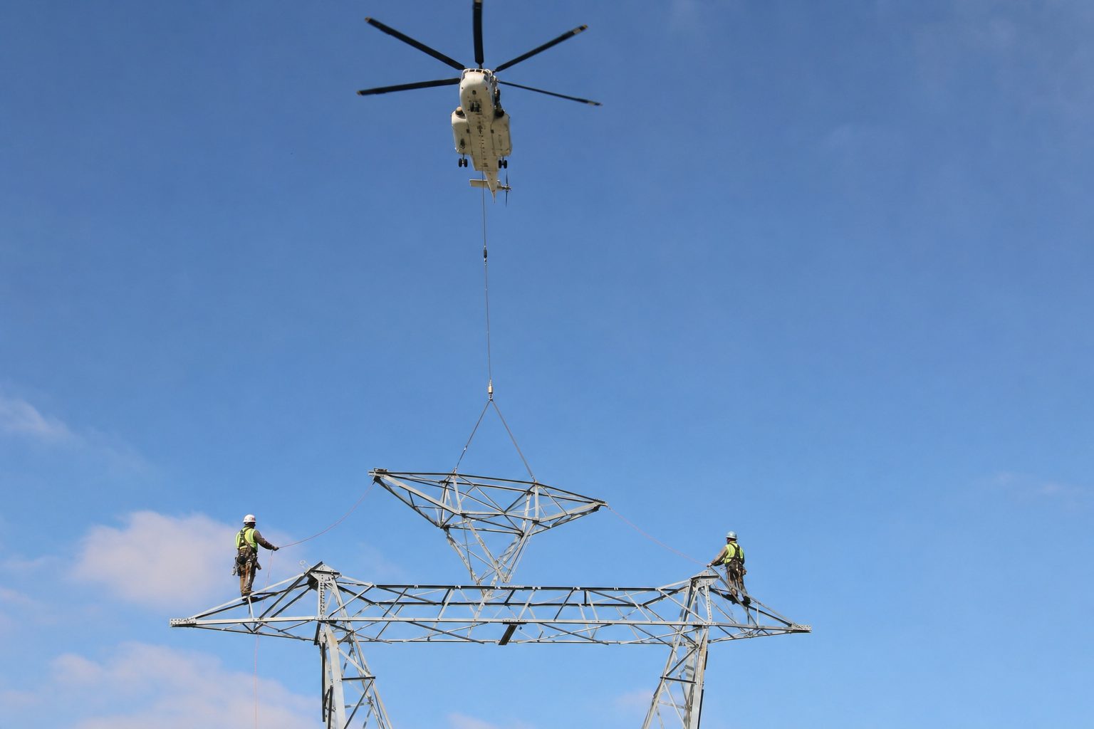 Helicopter on a longline lowering a galvanized lattice tower section onto a partially erected transmission tower while two ironworkers tend tag lines