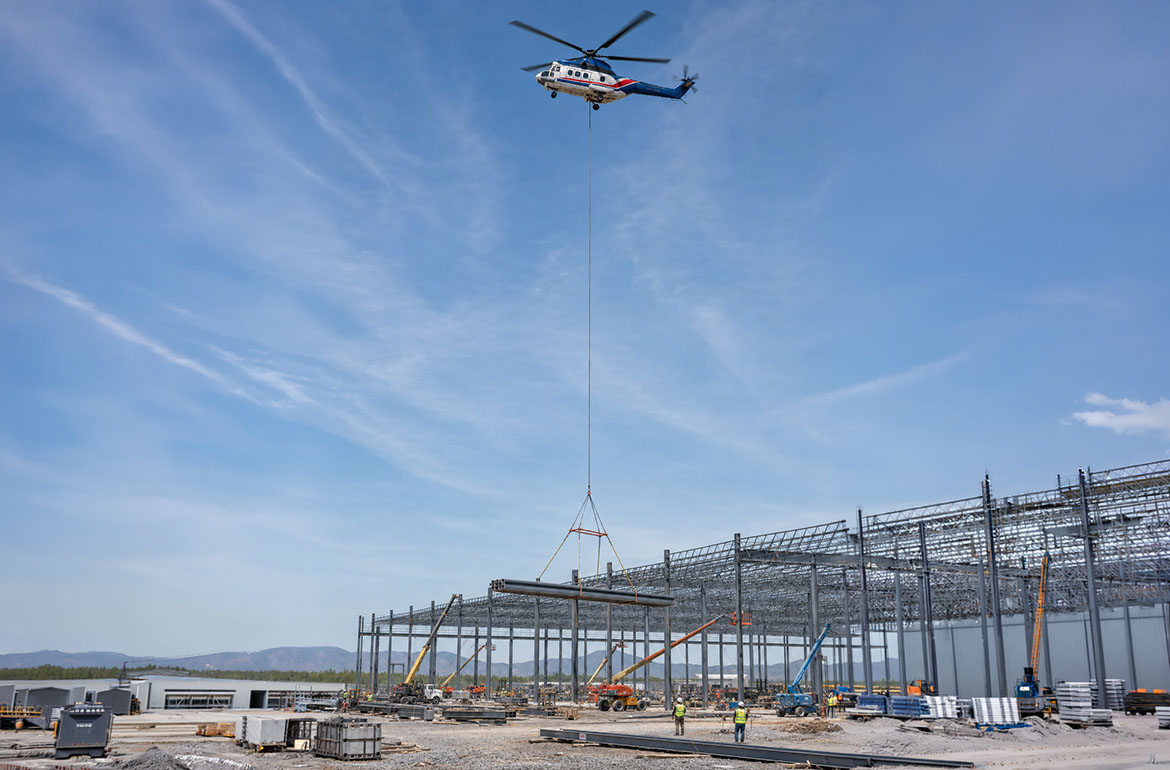 Heavy twin helicopter lifting a steel girder over an active commercial construction site