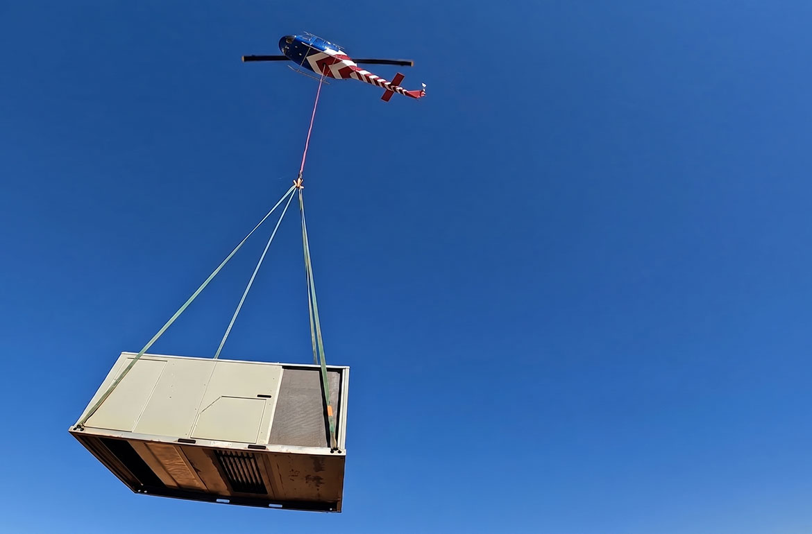 Light utility helicopter carrying a tan HVAC condenser on a longline against blue sky